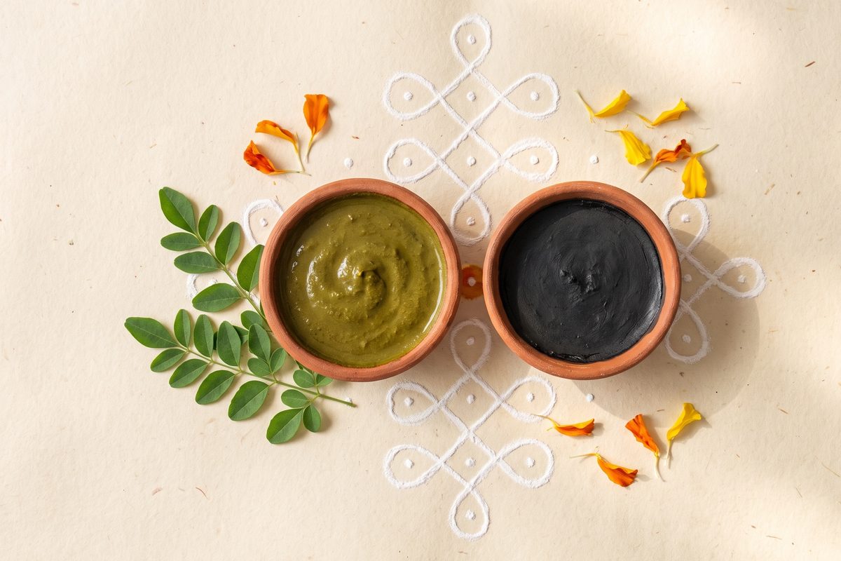 Gouache still life from above: two small terracotta bowls, one of green henna paste and one of flat black paste, among henna sprigs, marigold petals and a white rice-flour kolam pattern; a generated illustration, not a photograph
