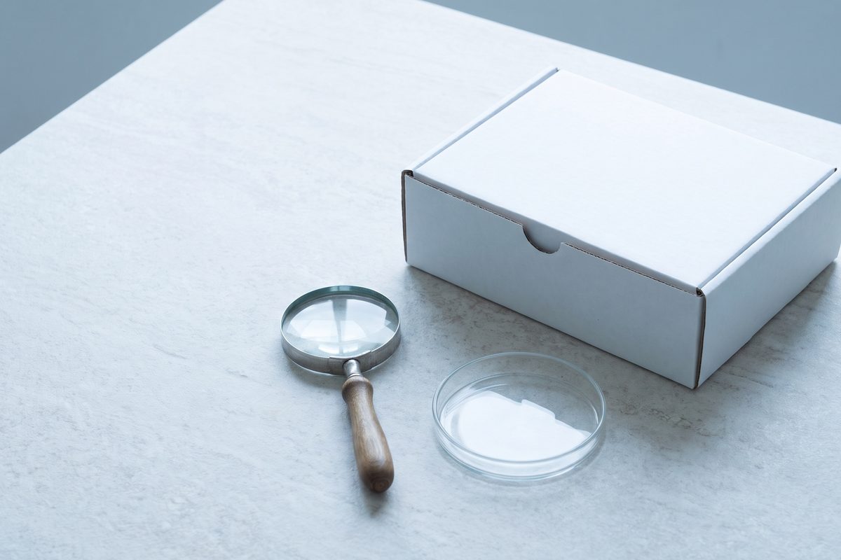 A still life of a closed, blank white cardboard box and a small empty glass dish beside a magnifying glass on a pale stone table in cool daylight; a generated image, not a photograph of a real scene (PROVISIONAL, check against the final image)