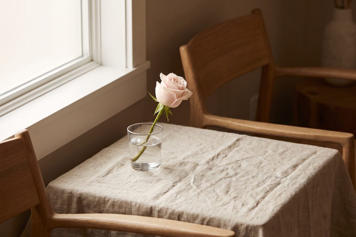 A folded pink silk ribbon on pale linen beside a glass of water in soft daylight, a still life with no people and no medical items in view