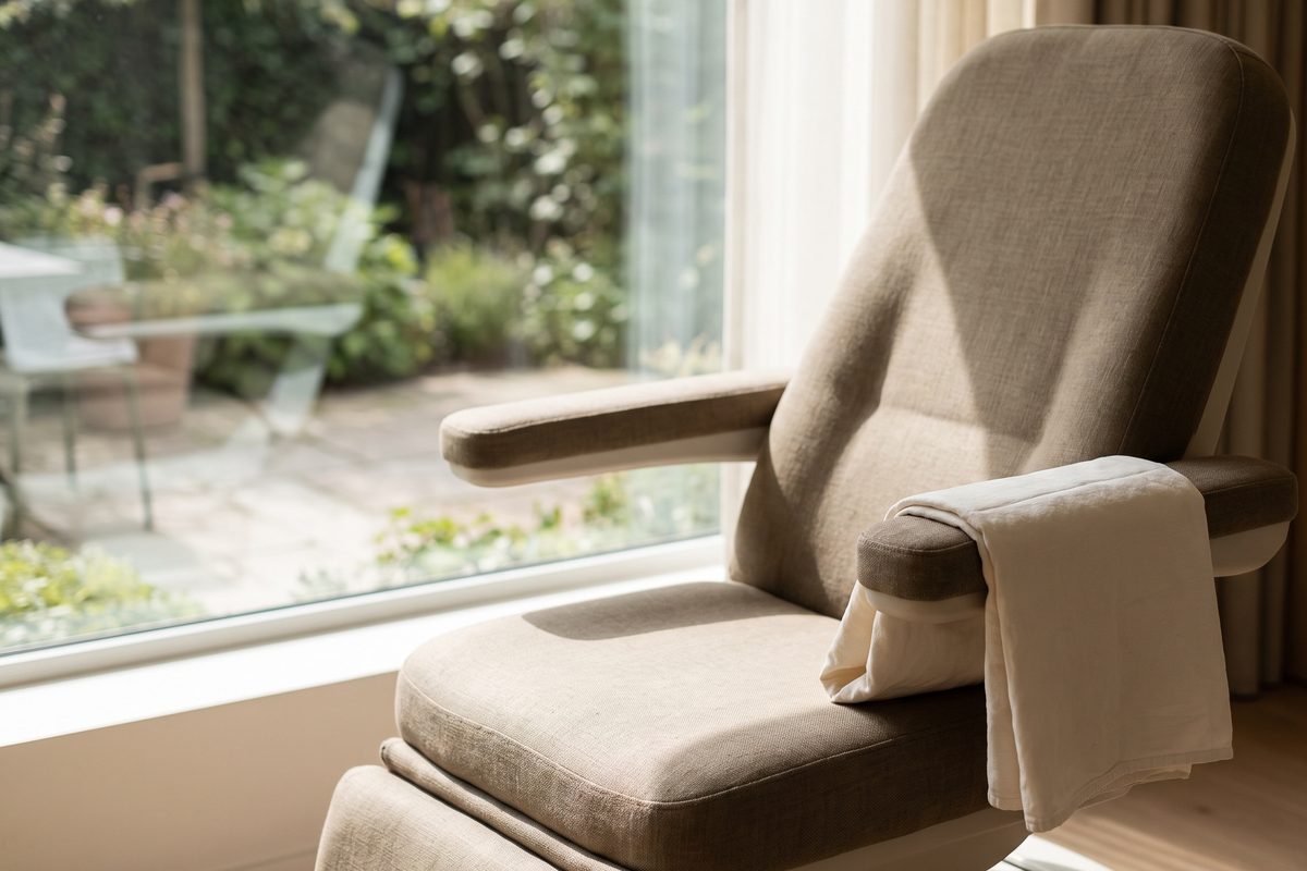 An empty examination chair beside a window in late morning daylight, a folded cotton drape on the armrest, no patient and no treatment tray present