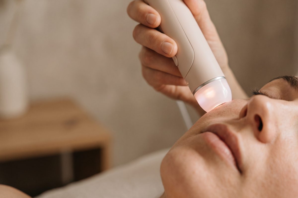 A pale treatment handpiece with a soft pink light held near the cheek of a relaxed woman lying on a couch, warm daylight, no text