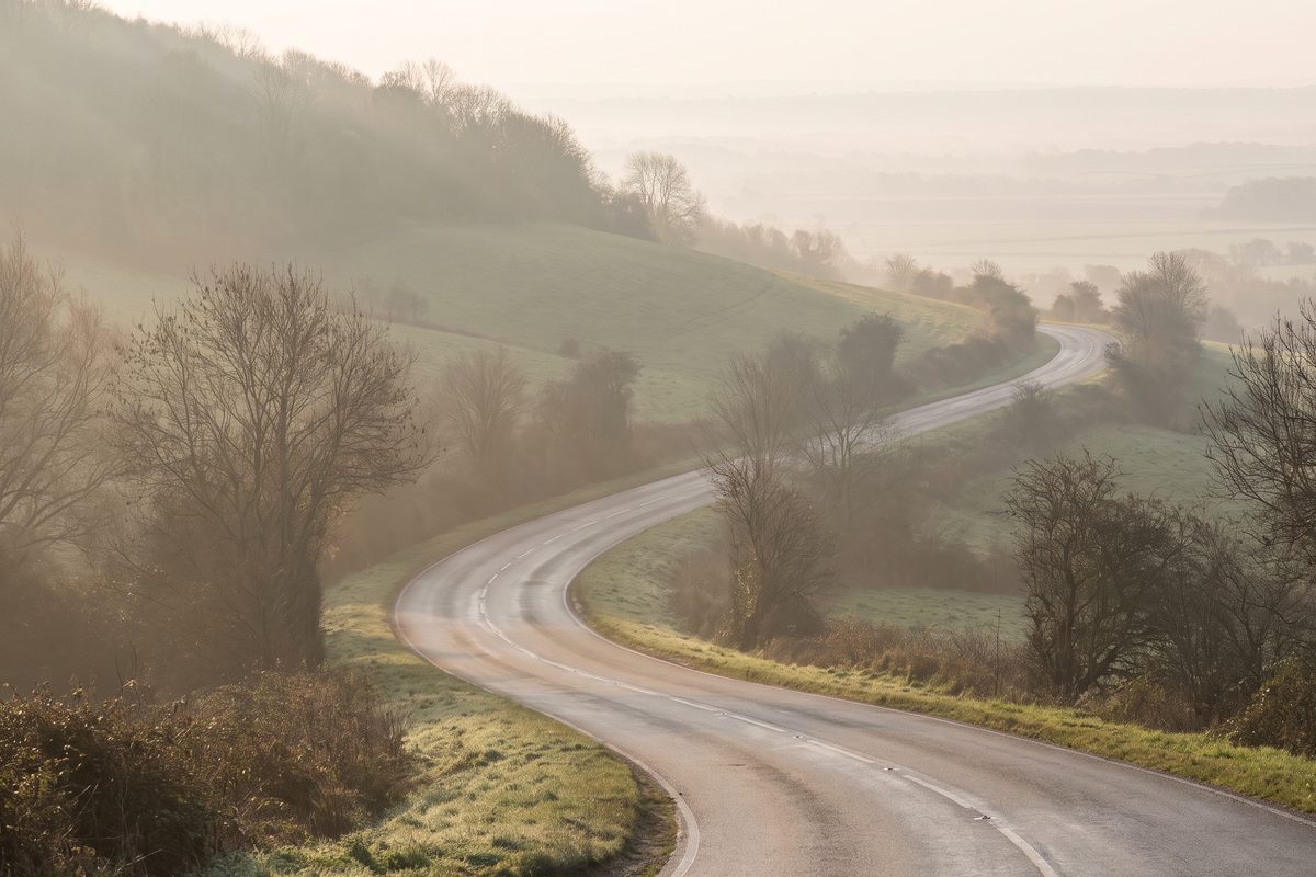 A hillside road curving downhill through morning mist, the gradient easing as it flattens towards level ground, with no people or vehicles present