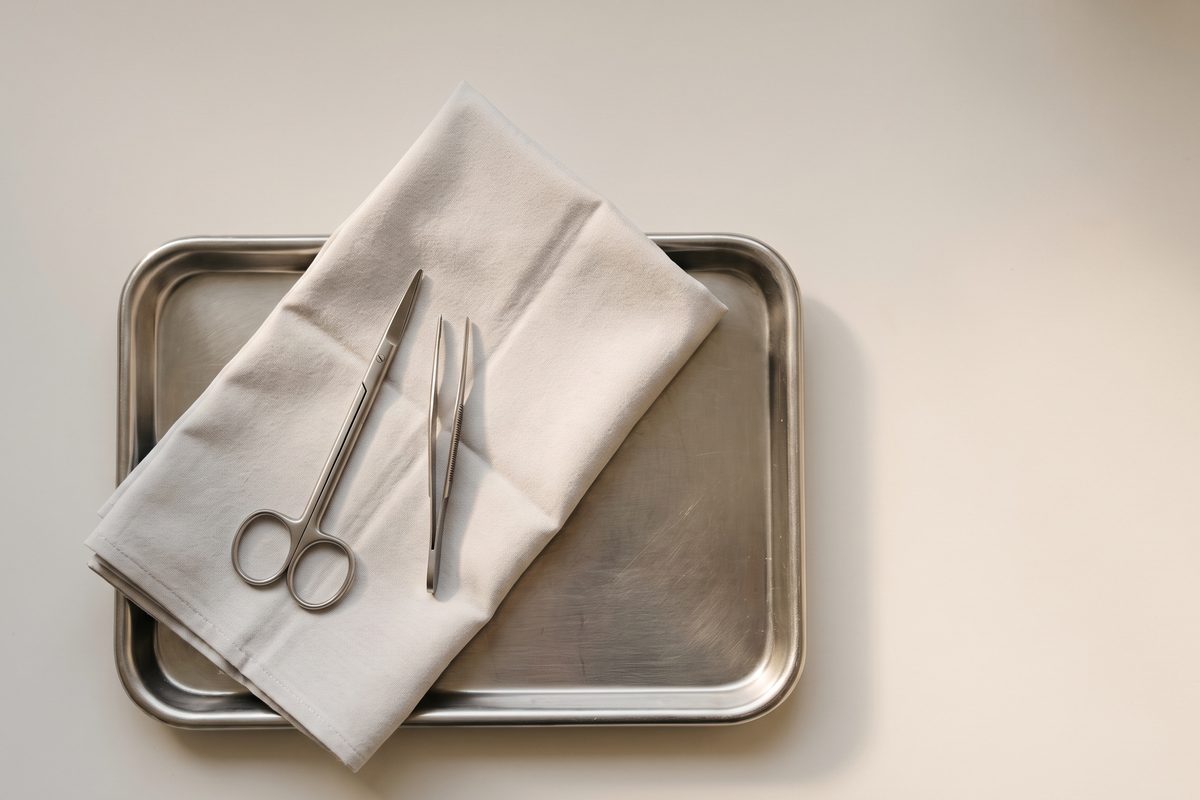 A pair of fine surgical scissors and a folded sterile drape resting on a stainless steel tray in flat daylight, no patient, no face and no procedure in frame