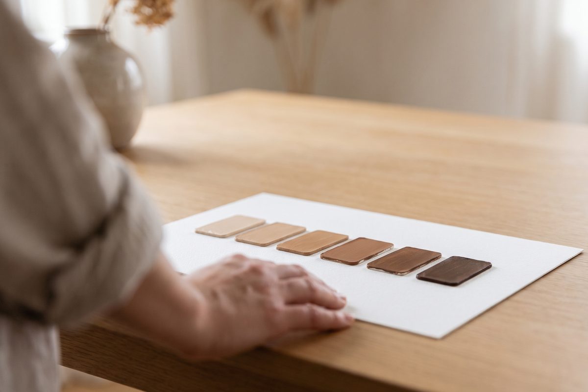 Six unlabelled paint swatches in graded shades of brown laid in a row on white card in flat daylight, no skin, no people and no lettering in frame