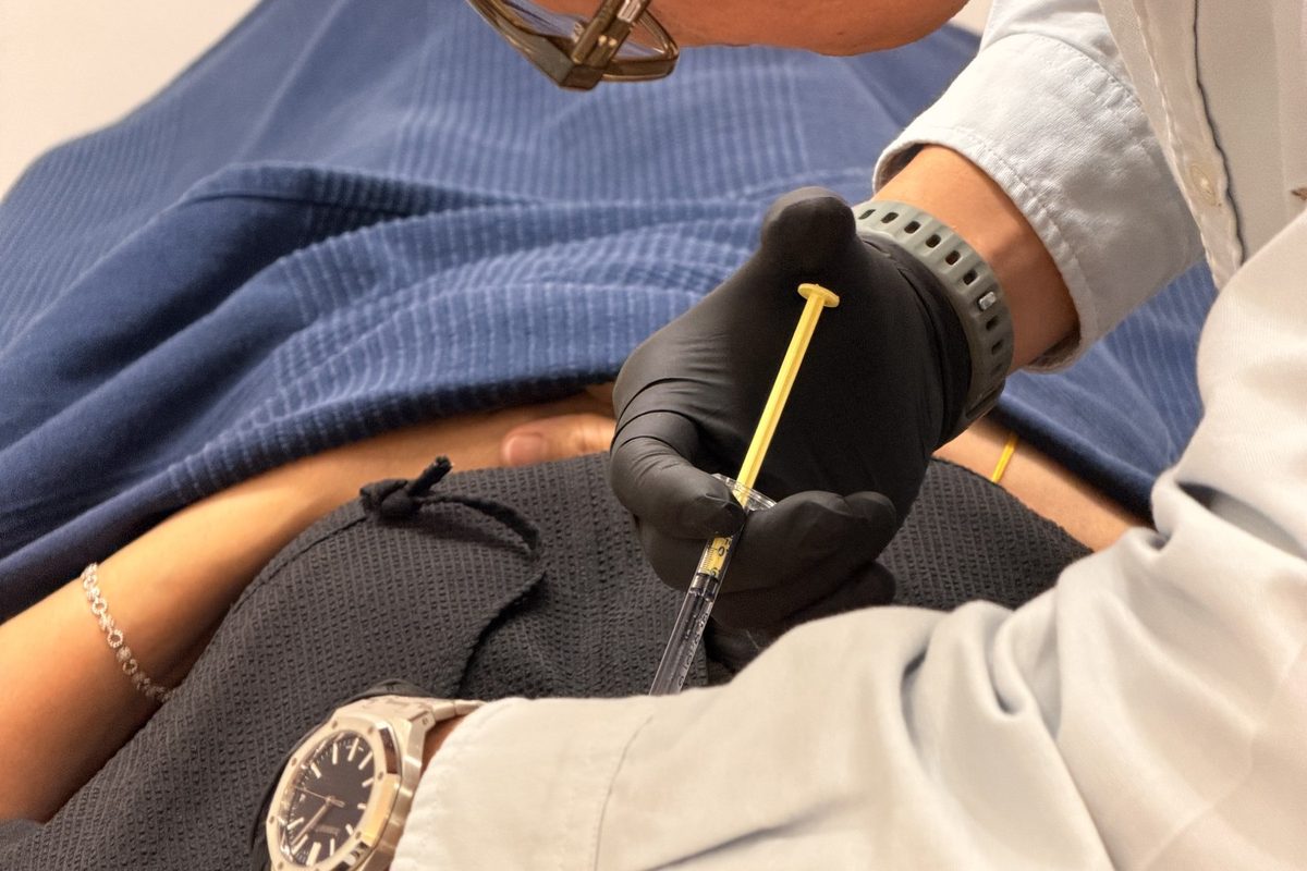 Dr Ong Jin Khang's gloved hands administering an injection into a patient's forearm in a clinic treatment room, documentary style
