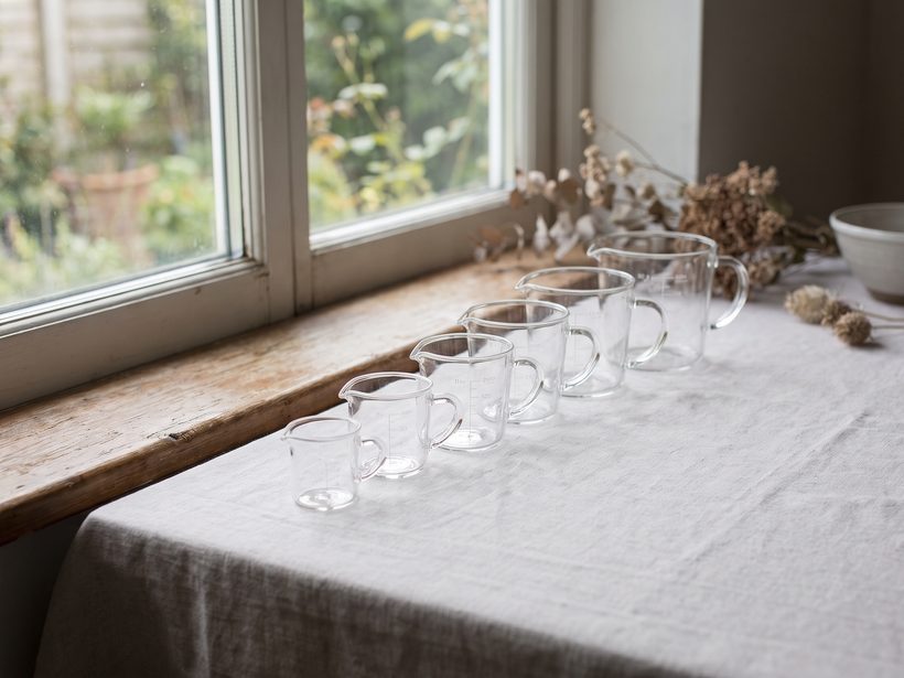 A row of six clear glass measuring jugs, from tiny to large, on a linen-covered table by a window