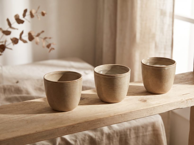 Three small ceramic cups in a row on a pale wooden shelf by a window, soft morning light, no person, no text