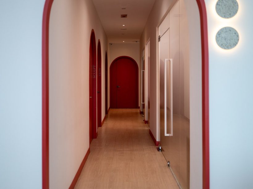 The Retreat Clinic corridor at rest, red arches receding toward a closed red door, nobody in frame