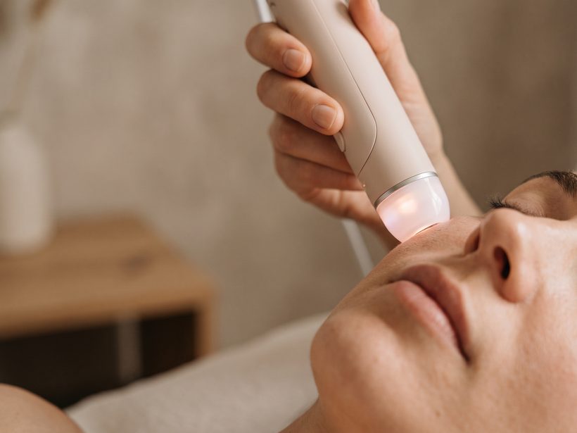 A pale treatment handpiece with a soft pink light held near the cheek of a relaxed woman lying on a couch, warm daylight, no text