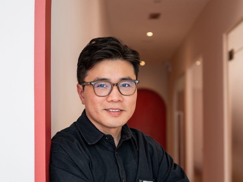 Dr Ong Jin Khang stands with arms crossed in the clinic's red-arch corridor, a calm, direct presence with no device or patient in frame