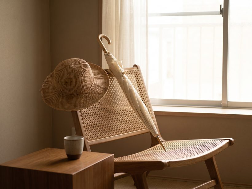 A straw sun hat and a closed cream parasol resting on a cane chair beside a sunlit window, a quiet still life with no people